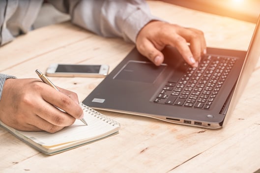 a picture with a laptop and the hands of a man, his left habd on the keyboard while the right hand is holding a pen and taking notes on a notebook and an iphone between the arms and in the space between his chest and the laptop sitting on the desk