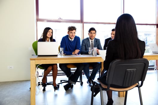 4 people sitting next to eachother on a table sitting in front of a woman with black hair sitting in a foldable chair with her back towards as and they 4 people looking to be in an interview